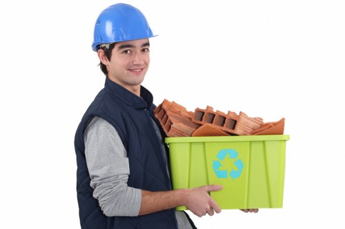 A young man wearing a blue safety helmet and a dark blue vest over a light grey long-sleeved shirt is smiling and holding a green recycling bin with a blue recycling symbol printed on its side. The bin is filled with red clay bricks, some of which have hollow sections, and appears to be made of durable plastic. He is standing against a plain white background, and the scene is professionally staged, likely for a rubbish removal service website. The image suggests a focus on construction or renovation debris clearance, fitting within the context of waste management services in or around Hoxton. The visual emphasizes careful handling of building materials and environmental responsibility associated with waste disposal or recycling. The setting is simple, with no additional environment or equipment visible, highlighting the worker's role in managing building waste in a professional manner.
