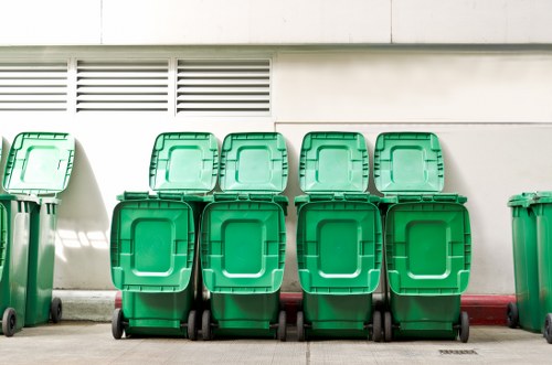A row of eight green wheeled garbage bins with closed lids, positioned on a paved surface in front of a white building with ventilation slats near the top. The bins are arranged closely together, with six placed side by side and two extra bins on their left, partially visible. The surface beneath the bins is a smooth concrete or tarmac, and a red line runs along the base of the wall behind them. The lighting suggests natural daylight, casting faint shadows on the ground and the building. This image visually represents waste collection or rubbish disposal services, relevant to Hoxton House Clearance and the local area.