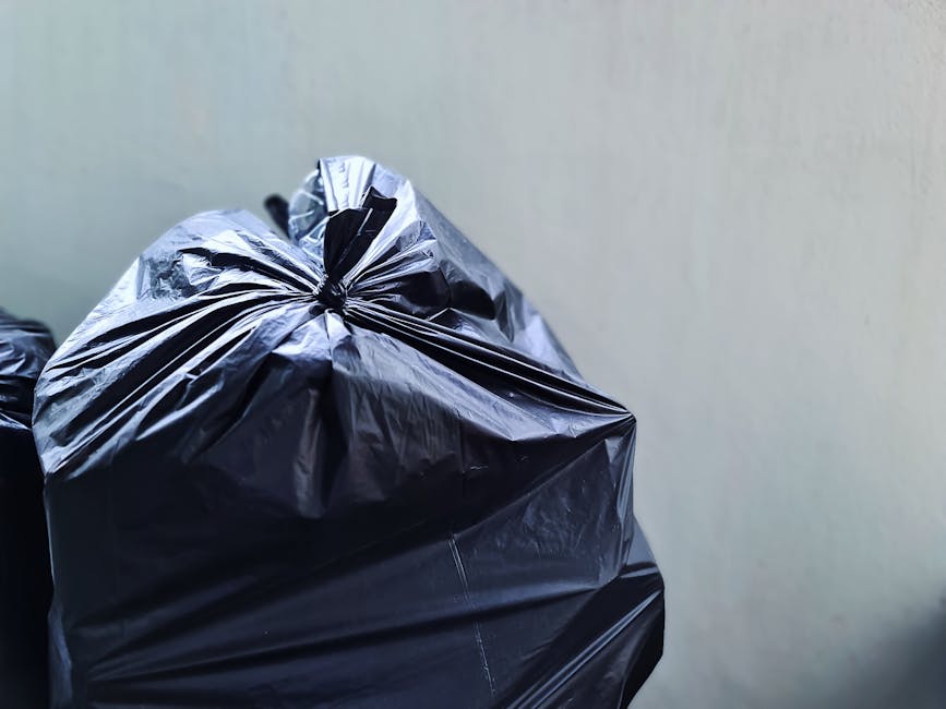 A close-up view of two black plastic rubbish bags tightly tied at the top, with one bag positioned slightly in front of the other. The bags have a shiny, crinkled surface, characteristic of standard r