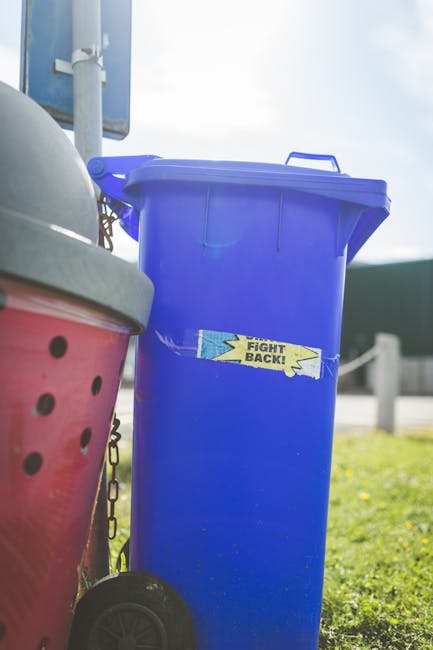 A green wheelie bin filled with several red and black bags of gravel or similar aggregate material, positioned outdoors on a paved surface next to a dark brick wall. The bin is placed on a wooden pall