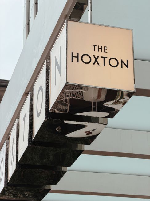 A narrow cobbled street within an urban setting, featuring a corner with a white brick building on the right displaying a street sign reading 'Hoxton Square'. To the left is a modern, multi-storey bui