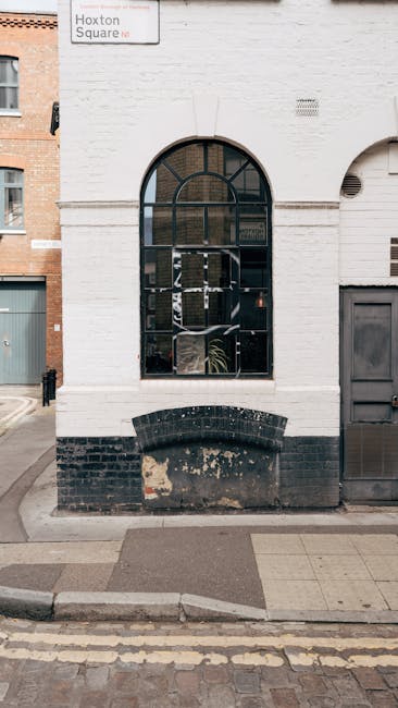 The image depicts the exterior of a white brick building with a large, arched window divided into multiple panes, some of which are obscured by reflections or objects inside. Below the window, there i